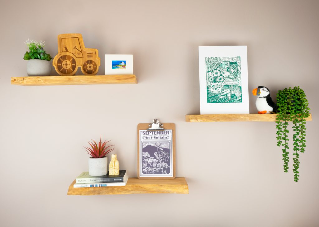 Three shelves on a wall, with various products and items including prints, a wooden tractor, a toy puffin and pot plants.